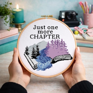 May include: A cross-stitch embroidery hoop featuring a landscape design with a waterfall, trees, and mountains in shades of blue, gray, and purple. The text "Just one more CHAPTER" is stitched above the landscape. The hoop is held by hands.