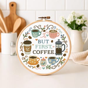 May include: A round, wooden embroidery hoop featuring a cross-stitch design with coffee-themed imagery. The design includes various coffee cups, a French press, and the text "BUT FIRST, COFFEE" in a light blue color. The color palette includes shades of brown, blue, pink, and yellow.