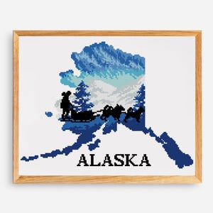 May include: Cross-stitch art of the Alaska state outline, featuring a dog sled team, mountains, and trees in shades of blue and white. The word "ALASKA" is stitched in black block letters. The artwork is framed in a light brown wooden frame.
