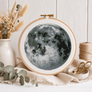 May include: A cross-stitch embroidery of the moon in a wooden hoop. The moon is rendered in grey and black, with detailed shading to represent the lunar surface. The hoop is displayed on a neutral-coloured fabric.