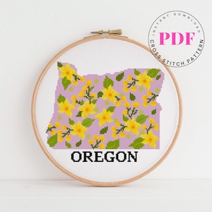 May include: Cross-stitch pattern of the state of Oregon, filled with yellow flowers and green leaves on a lavender background. The word "OREGON" is stitched in black at the bottom. The design is framed in a wooden embroidery hoop, with the text "INSTANT DOWNLOAD PDF CROSS STITCH PATTERN" in a circle.