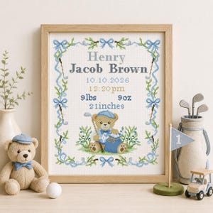 May include: A framed cross-stitch birth announcement featuring a teddy bear golfer, blue bows, and green foliage. The text reads Henry Jacob Brown, 10.10.2026, 12:20pm, 9lbs 9oz, and 21inches. The design is enclosed in a light wood frame.