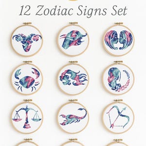 May include: Twelve embroidered zodiac signs displayed in wooden hoops. Each design, in blue, pink, and white, represents a different astrological sign. The text "12 Zodiac Signs Set" is above the hoops, with a gold zodiac wheel.