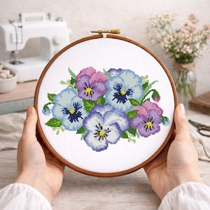 May include: A cross-stitch embroidery of pansies in shades of blue, purple, and pink, with green leaves. The floral design is set in a wooden embroidery hoop. The embroidery is held by a person.