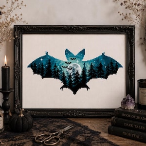 May include: A framed cross-stitch embroidery featuring a bat silhouette filled with a blue and black forest landscape, a full moon, and small stars. The bat is centered on a white fabric background within a black ornate rectangular frame.
