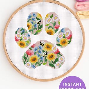 May include: A cross-stitch paw print design, featuring flowers and bees, displayed in a wooden embroidery hoop. The design incorporates sunflowers, blue flowers, and pink hues. A purple circle with the text "INSTANT DOWNLOAD PDF" is in the lower right.