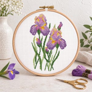May include: A circular wooden embroidery hoop containing a cross-stitch pattern of two purple and yellow iris flowers with green stems and leaves on white fabric. The design features detailed pixelated floral motifs in shades of violet and gold.
