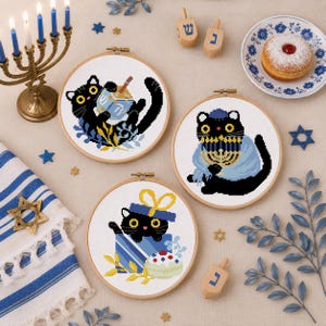 May include: Three cross-stitch hoops featuring black cats celebrating Hanukkah. One cat is holding a bowl, another is wearing a menorah, and the third is in a gift box. The scene includes a menorah, dreidels, and a sufganiyah.