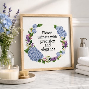 Puede incluir: Un marco de madera cuadrado que contiene un bordado de punto de cruz de una corona floral hecha de hortensias azules, lavanda morada y pequeñas flores blancas. El texto central dice: Please urinate with precision and elegance.