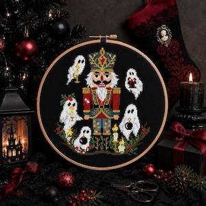 May include: A circular wooden embroidery hoop containing a cross-stitch design on black fabric. The pattern features a central nutcracker figure in red, blue, and gold, surrounded by five white ghosts holding holiday items like candles and ornaments.