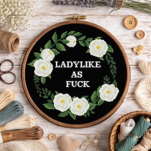 Peut inclure: Broderie au point de croix dans un cercle en bois. Le tissu noir présente une couronne florale avec des roses blanches et des feuilles vertes. Le texte "LADYLIKE AS FUCK" est centré dans la couronne. Des fournitures d'artisanat, dont du fil et des ciseaux, entourent le cercle.