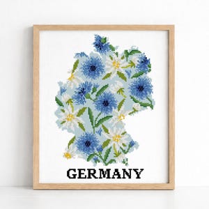 Può includere: Un'opera d'arte a punto croce incorniciata raffigurante la mappa della Germania riempita di fiori blu e bianchi e foglie verdi. La parola "GERMANY" è stampata in lettere nere in basso. L'opera è ambientata su uno sfondo bianco.