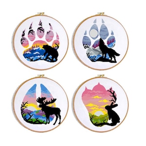 May include: Four circular embroidery hoops with cross-stitch designs depicting nature scenes. Each features a sunset within a paw print shape, with silhouettes of a bear, wolf, moose, and rabbit. The hoops have wooden frames.