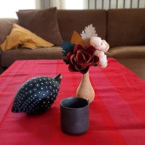 May include: A black and white speckled ceramic bird figurine sits on a red and white striped tablecloth next to a black ceramic mug and a wooden vase with a red paper flower and two white paper flowers.