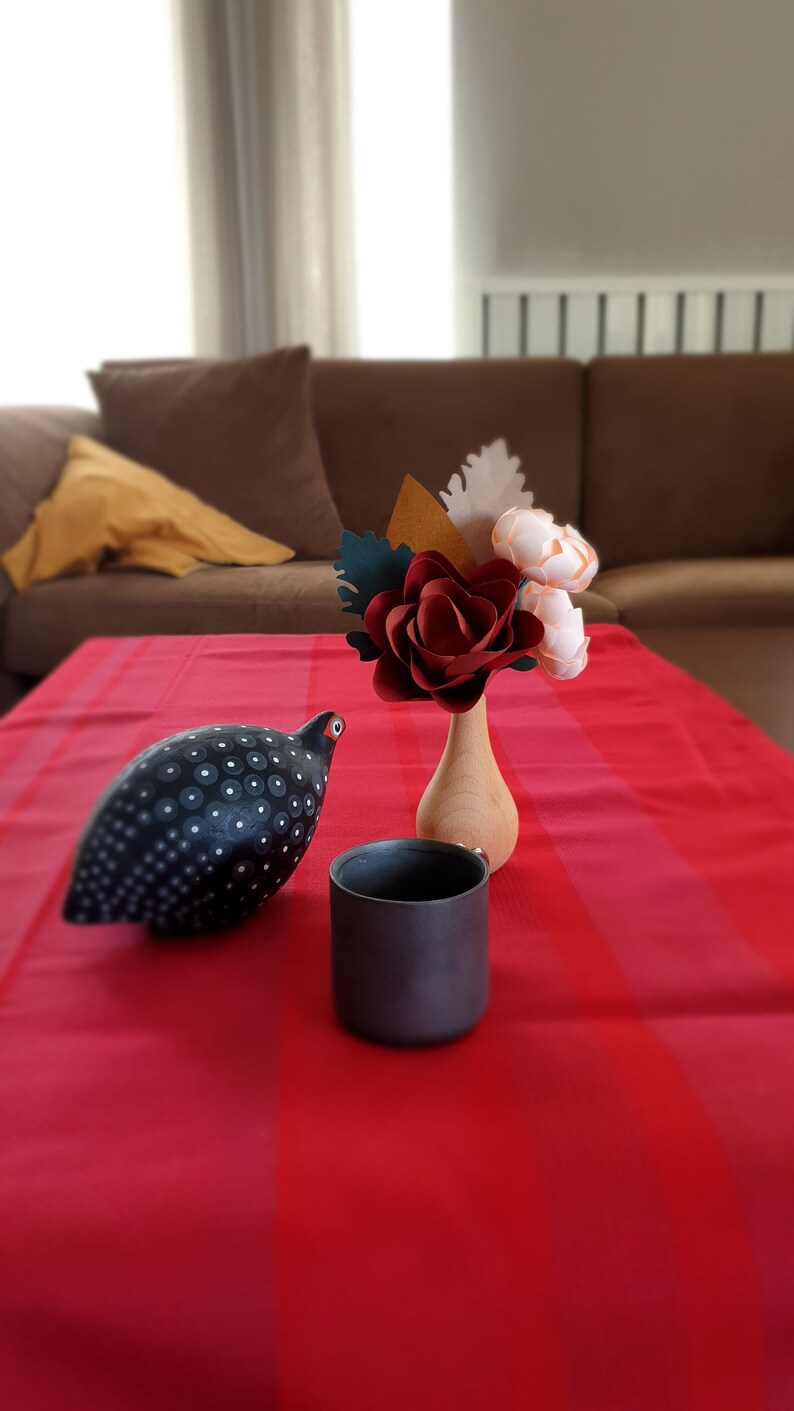 May include: A black and white speckled ceramic bird figurine sits on a red and white striped tablecloth next to a black ceramic mug and a wooden vase with a red paper flower and two white paper flowers.
