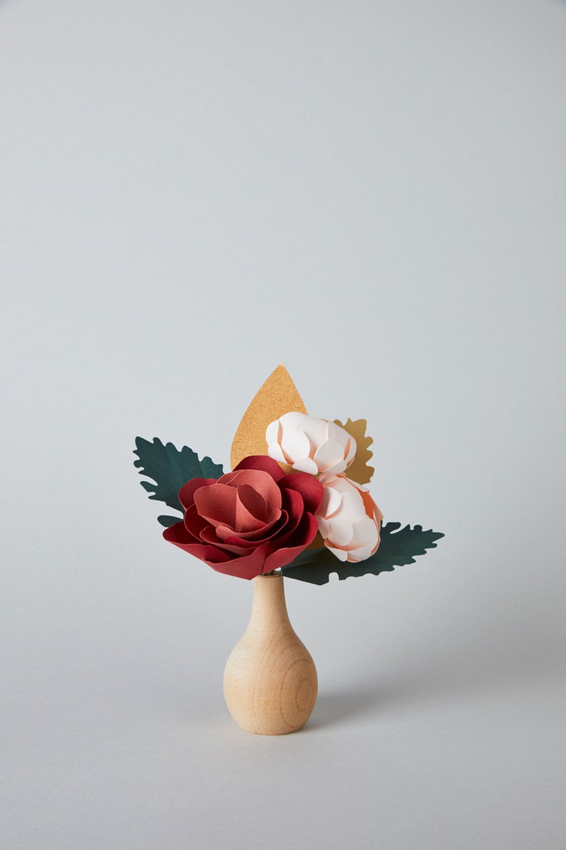 May include: A small wooden vase with a paper flower arrangement. The flowers are red, pink, and white, and the leaves are green and gold.