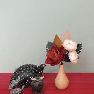 May include: A black and white spotted ceramic bird figurine, a brown ceramic bull figurine, and two smaller black and white spotted ceramic bird figurines on a red striped surface. A vase with a red, pink, and brown paper flower arrangement sits on the surface.