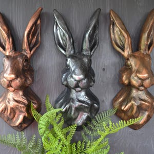 May include: Three rabbit head sculptures mounted on a wall. The rabbit heads are made of a textured material and are painted in copper, black, and bronze.