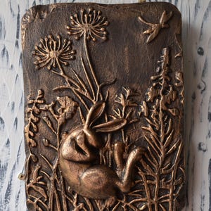 May include: A bronze-colored wall sculpture depicting a rabbit resting in a field of tall grasses and flowers. The sculpture is detailed with raised textures and a rustic finish.