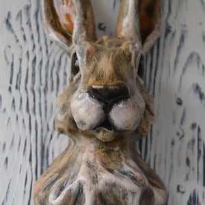 May include: A ceramic wall-mounted sculpture of a hare head and neck. The piece features a textured, hand-painted finish in shades of brown, tan, and white, with glossy accents on the ears and nose, capturing the animal's alert facial expression.