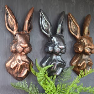 May include: Three decorative rabbit head wall sculptures in different metallic finishes: copper, silver, and gold. The sculptures are mounted on a dark grey wall.