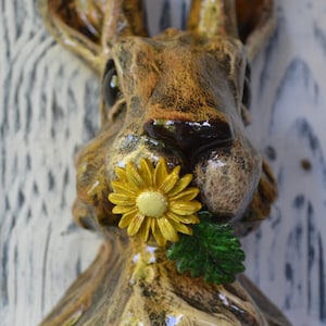 May include: A ceramic wall-mounted hare sculpture with a mottled brown and tan finish. The hare has long, upright ears and is depicted holding a bright yellow daisy flower with a small green leaf in its mouth, featuring a glossy glazed texture.