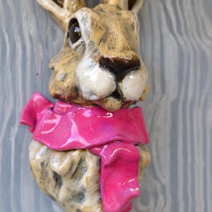 May include: A ceramic hare sculpture with a tan and brown face, long ears, and a bright pink scarf. The sculpture is mounted on a light blue and grey wooden background. The hare's features are detailed, with a focus on the eyes and nose.
