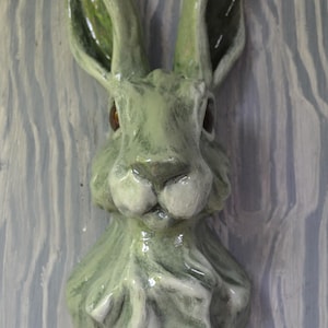 May include: A ceramic wall-mounted sculpture of a rabbit head and neck. The piece features a glossy, light sage green glaze with darker green shading in the crevices and textured fur details. The rabbit has upright ears and dark brown eyes.
