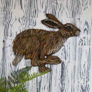 May include: A detailed, brown wooden hare sculpture in a running pose. The sculpture has textured fur and is mounted on a white and grey wooden background. The hare is a decorative item, possibly for a wall.