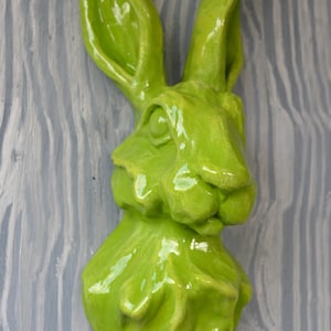 May include: A vibrant, glossy green rabbit head sculpture. The sculpture is mounted on a light gray, wood-grain textured background. The rabbit has long ears and a detailed face, creating a whimsical decorative piece.