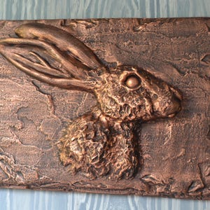 May include: A bronze-toned rectangular wall plaque with a detailed relief of a hare's head and upper body. The hare's long ears are prominent, and the textured surface adds depth. The artwork is set against a textured background.