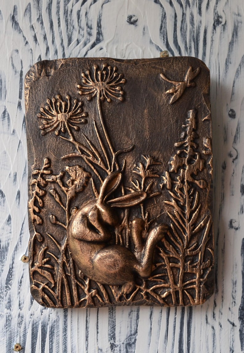 May include: A bronze-colored wall sculpture depicting a rabbit resting in a field of tall grasses and flowers. The sculpture is detailed with raised textures and a rustic finish.