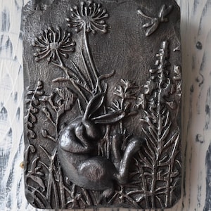 May include: A silver metal wall sculpture depicting a sleeping rabbit surrounded by stylized plants and a dragonfly.