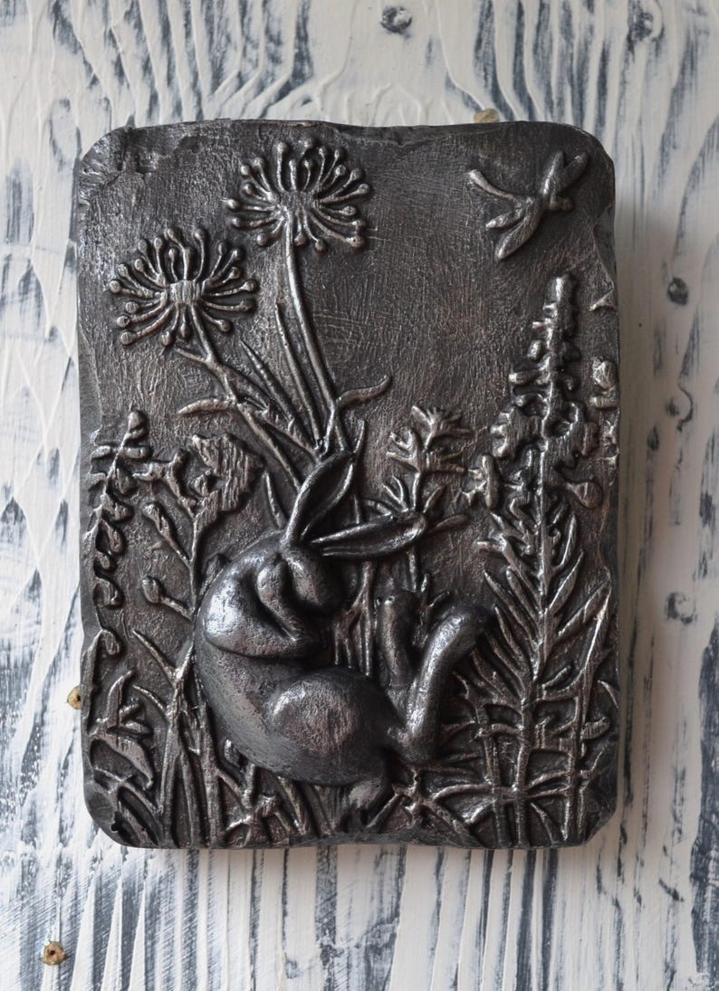 May include: A silver metal wall sculpture depicting a sleeping rabbit surrounded by stylized plants and a dragonfly.