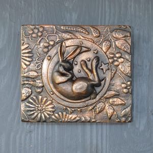 May include: A copper-colored wall hanging with a raised design of a rabbit sleeping in a crescent moon. The moon is surrounded by a floral design with leaves and berries.
