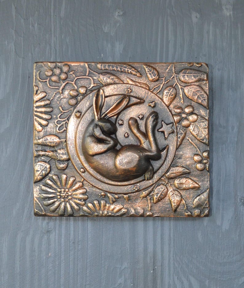 May include: A copper-colored wall hanging with a raised design of a rabbit sleeping in a crescent moon. The moon is surrounded by a floral design with leaves and berries.