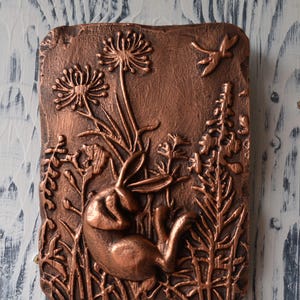 May include: A copper-colored wall sculpture depicting a rabbit resting in a field of flowers and grasses. The sculpture is detailed with a dragonfly in flight.