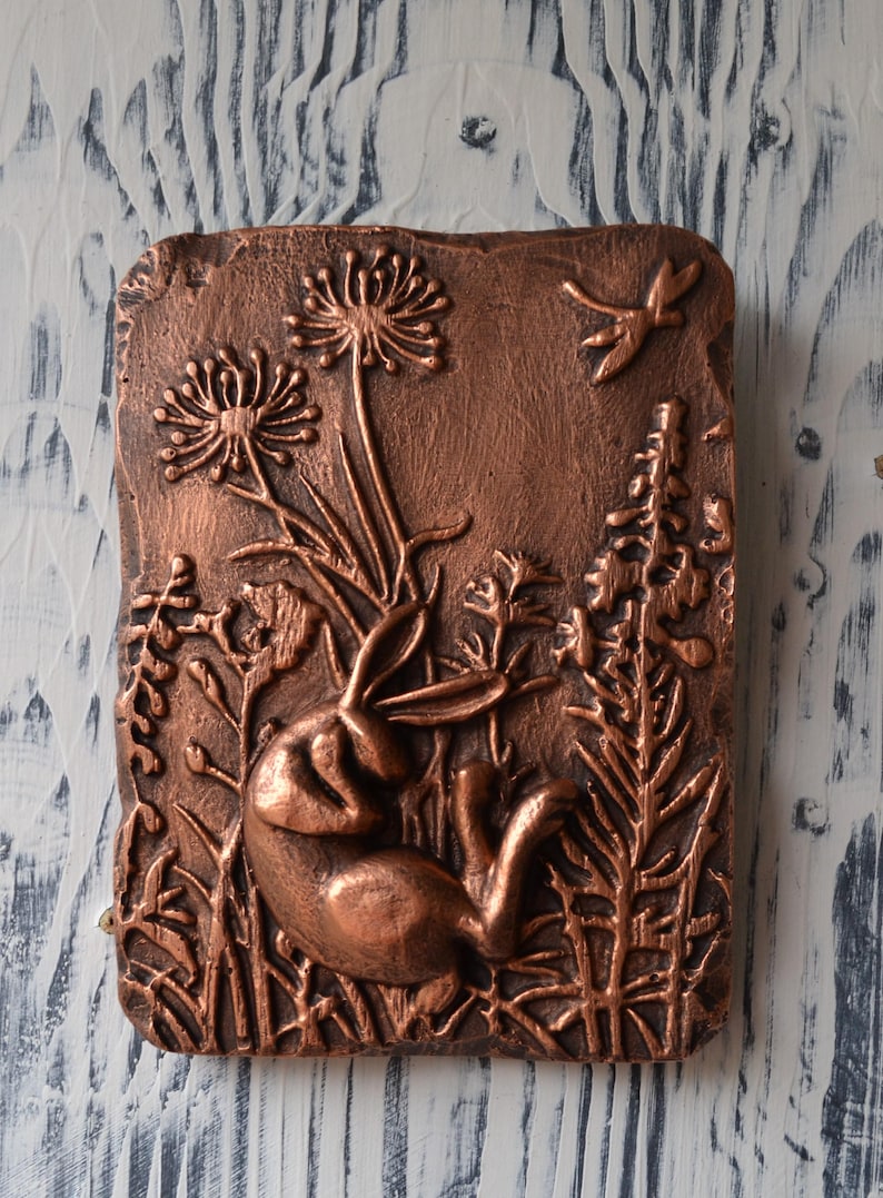 May include: A copper-colored wall sculpture depicting a rabbit resting in a field of flowers and grasses. The sculpture is detailed with a dragonfly in flight.