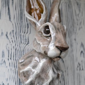 Peut inclure: Sculpture en céramique d'une tête de lièvre avec un glaçage brun et blanc. Le lièvre a de grandes oreilles et un visage détaillé.