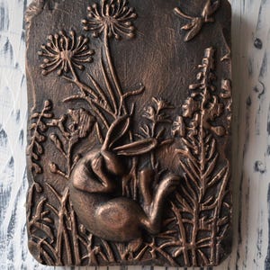 May include: A copper-colored wall plaque with a raised relief design of a rabbit in a field of flowers and a dragonfly. The plaque has a rustic, textured surface.