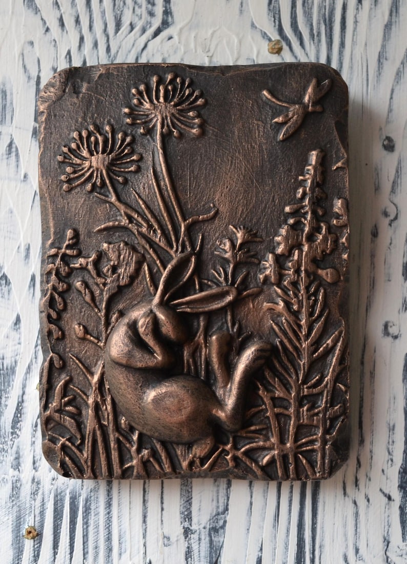 May include: A copper-colored wall plaque with a raised relief design of a rabbit in a field of flowers and a dragonfly. The plaque has a rustic, textured surface.