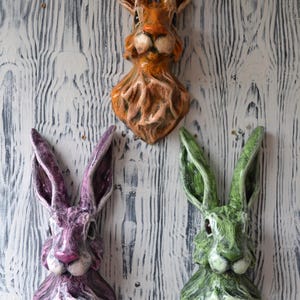 May include: Three decorative rabbit head wall hangings in orange, purple, and green. The rabbit sculptures have long ears and detailed facial features. The background is a white-washed wood grain.