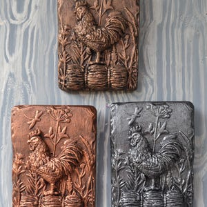 May include: Three rectangular wall plaques with a rooster design. The plaques are in bronze, copper, and silver tones. Each plaque features a detailed relief of a rooster standing on stacked barrels, surrounded by foliage.