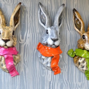 May include: Three ceramic rabbit head wall decorations. One is tan with a pink scarf, one is gray with an orange scarf, and one is tan with a green scarf. The rabbit heads are mounted on a light gray wooden background.