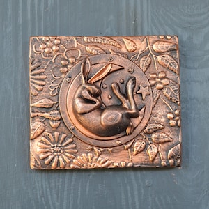 May include: A copper-colored wall hanging with a 3D sculpted design of a rabbit sleeping on a crescent moon. The moon is surrounded by a floral design with leaves and berries.
