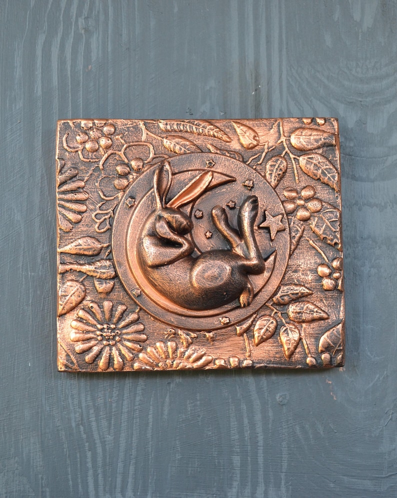 May include: A copper-colored wall hanging with a 3D sculpted design of a rabbit sleeping on a crescent moon. The moon is surrounded by a floral design with leaves and berries.