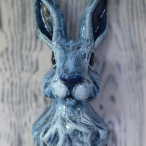 May include: A ceramic wall-mounted sculpture of a rabbit head featuring a glossy, mottled blue and white glaze. The rabbit has long, upright ears, dark eyes, and a textured surface that highlights the facial features and fur details in relief.