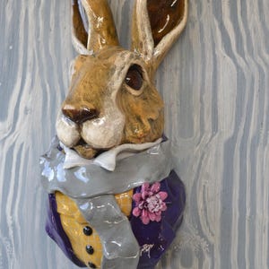 May include: A ceramic wall sculpture of a brown rabbit head and torso. The rabbit wears a purple jacket over a yellow vest with black buttons, a white collar, a grey scarf, and a pink flower pinned to the lapel. The finish is glossy and smooth.