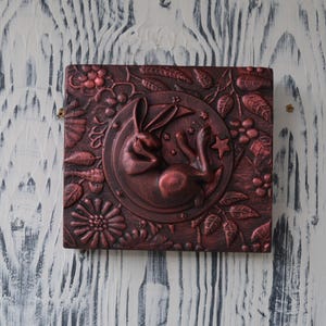 May include: A square, red-brown, wooden box with a raised, sculpted design of a rabbit sleeping on a crescent moon. The moon is surrounded by leaves, flowers, and stars.