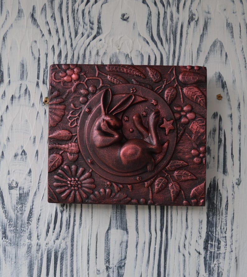 May include: A square, red-brown, wooden box with a raised, sculpted design of a rabbit sleeping on a crescent moon. The moon is surrounded by leaves, flowers, and stars.